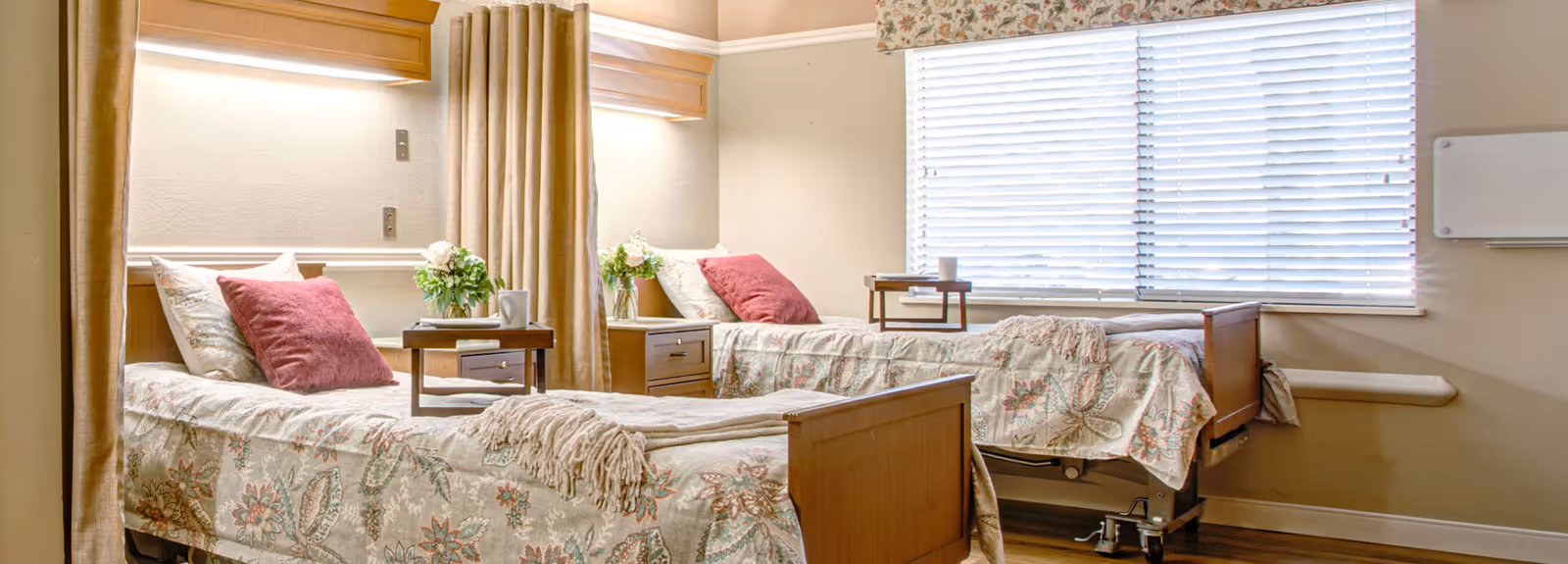 A bright and clean room with two hospital-style beds separated by a curtain. Each bed has floral-patterned bedding, a pink pillow, and a small wooden bedside table with a vase of flowers and a cup. There is a large window with white blinds letting in natural light.