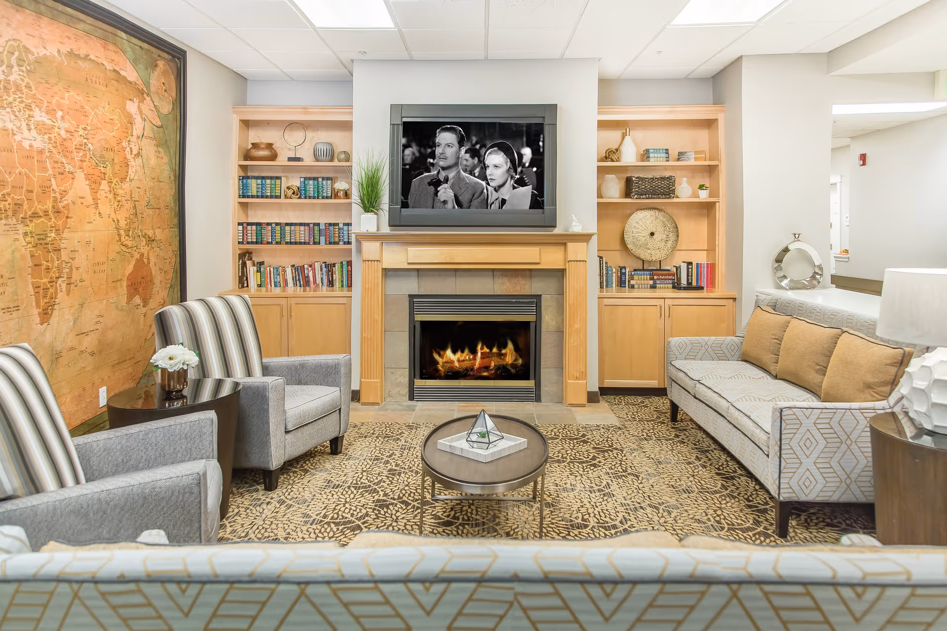 A cozy living room with a fireplace in the center, flanked by wooden built-in bookshelves filled with books and decorative items. Above the fireplace is a TV showing a black and white movie scene. The room has a patterned area rug, two gray armchairs with striped cushions on the left, and a gray sofa with brown pillows on the right. A round coffee table with a decorative item sits in the middle. On the left wall, there is a large vintage-style world map.
