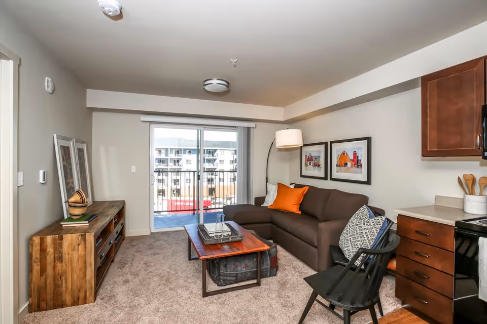 A cozy living room area with a brown sectional sofa adorned with orange and patterned pillows, a wooden coffee table with a tray and glassware, a black chair, and a wooden sideboard with decorative items. The room has beige carpet, light-colored walls, two framed pictures of barns on the wall, and a sliding glass door leading to a balcony with a view of neighboring buildings. Part of a kitchen with wooden cabinets and countertop is visible on the right.
