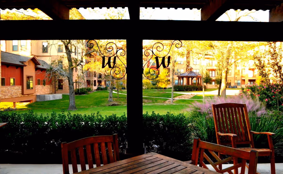 View from a covered patio with wooden chairs and a table looking out onto a green courtyard with trees, bushes, and a gazebo. Residential buildings surround the courtyard, and decorative wrought iron elements with the letter 'W' hang from the patio roof.
