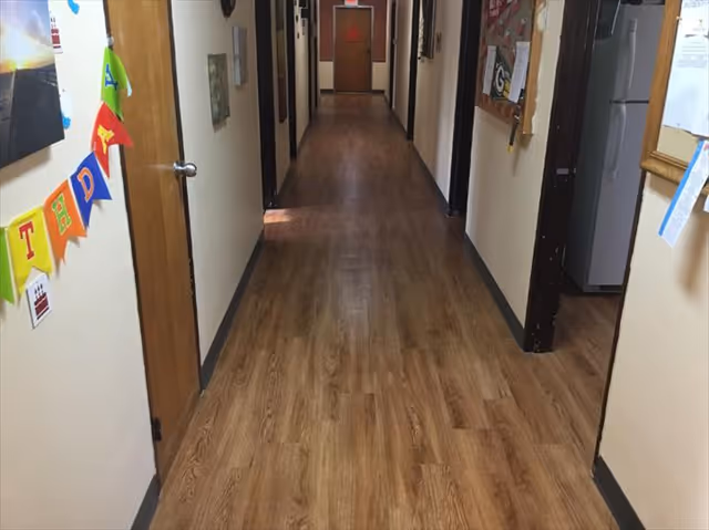 A long indoor hallway with wooden flooring and beige walls. Several closed wooden doors line both sides of the hallway. On the left wall, colorful triangular flags spell out the word 'THE DAY'. On the right side, a bulletin board and a refrigerator are visible in an open doorway.