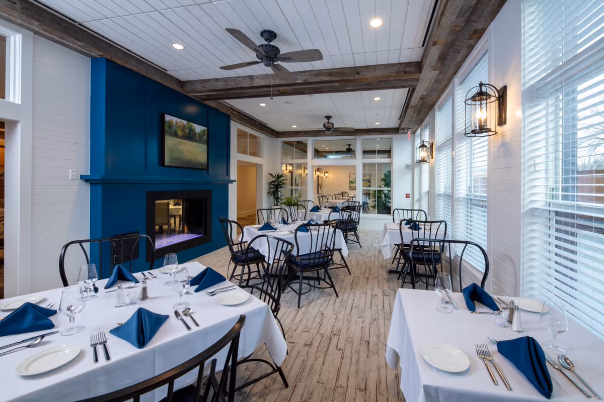 A bright dining room with white walls and large windows with blinds letting in natural light. The room features several tables covered with white tablecloths, each set with plates, silverware, wine glasses, and blue folded napkins. Black metal chairs surround the tables. A blue fireplace with a landscape painting above it is on one wall. Ceiling fans and rustic wooden beams adorn the ceiling, and wall-mounted lantern-style lights provide additional lighting.