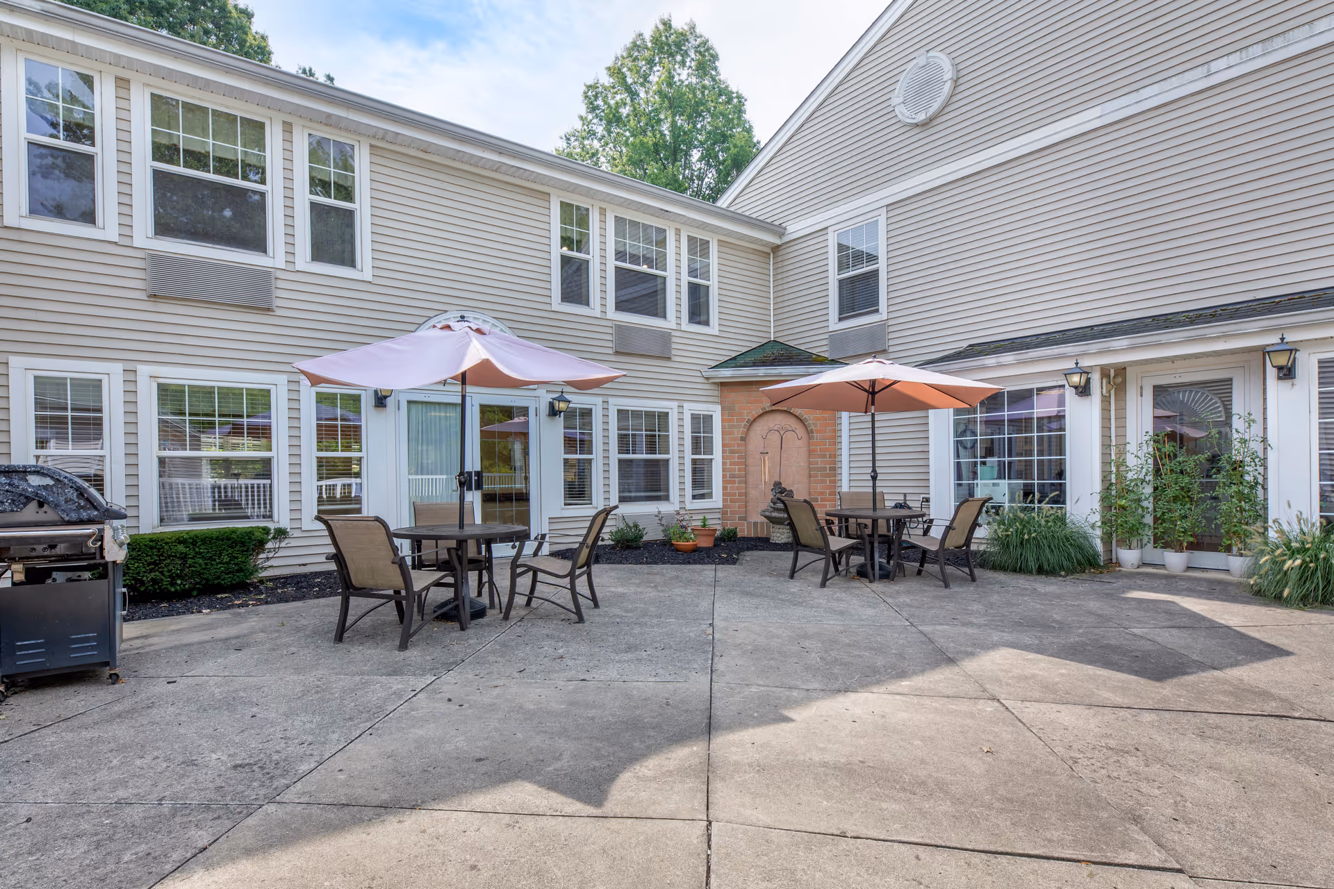 Outdoor patio area at Brookdale Wickliffe with two round tables, each shaded by a pink umbrella and surrounded by chairs. The patio is paved with concrete and adjacent to a beige building with multiple windows and glass doors. There is a barbecue grill on the left side and some potted plants near the building.