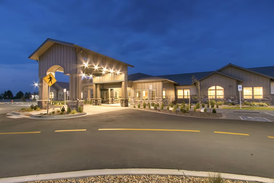 Exterior view of a senior living facility building at dusk with lights illuminating the entrance area. The building has a covered drop-off area with stone pillars and a pedestrian crossing sign nearby. The sky is dark blue, and the parking lot is visible in the foreground.
