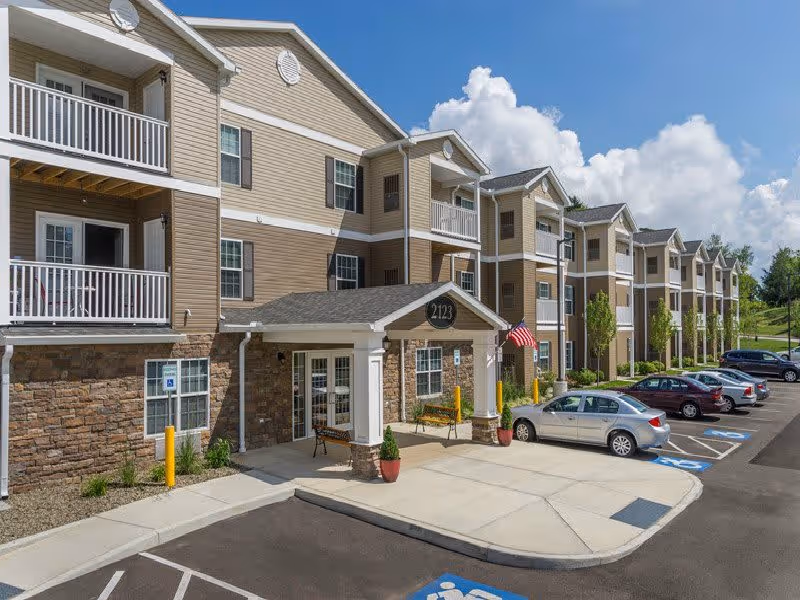 Exterior view of a multi-story residential building with beige siding and stone accents, featuring balconies and a covered entrance with the number 2123. Several cars are parked in front, including handicapped parking spaces, under a partly cloudy blue sky.