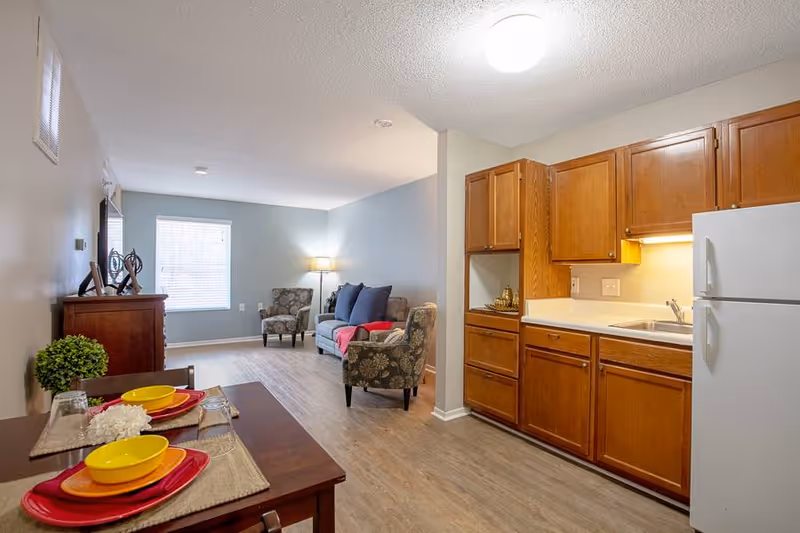 Interior view of a living space in Elison Assisted Living of Oxford showing a small kitchen area with wooden cabinets, a white refrigerator, and a sink. Adjacent to the kitchen is a dining table set with yellow bowls, red plates, and placemats. In the background, there is a living room area with a sofa, two patterned armchairs, a floor lamp, and a window letting in natural light.