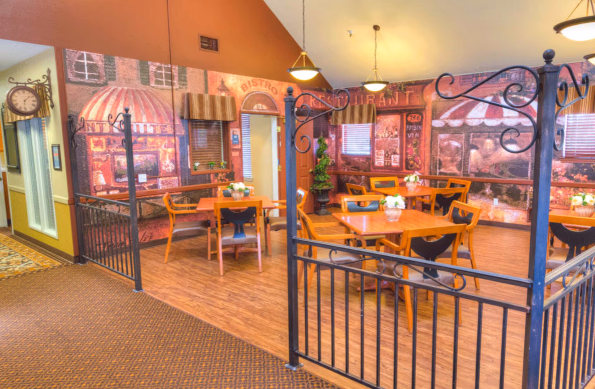 A cozy dining area in a senior living facility with wooden tables and chairs, decorated with small flower arrangements. The walls have murals depicting a quaint European street scene with storefronts and awnings. The space is warmly lit with hanging pendant lights and enclosed by decorative black metal railings.