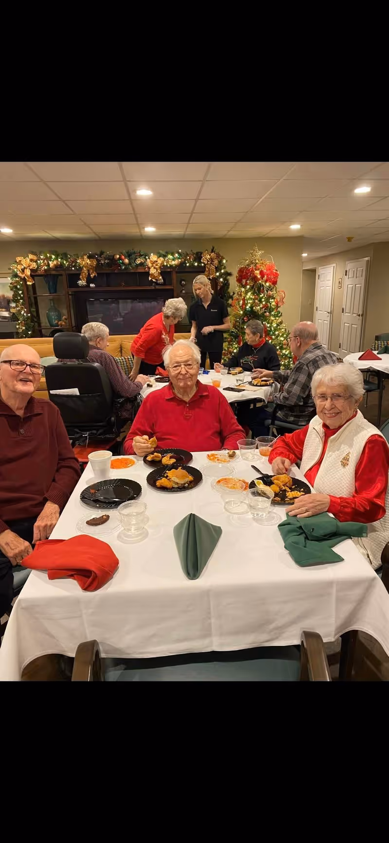 A group of elderly people sitting around a table enjoying a meal together in a festive room decorated with Christmas garlands and a Christmas tree. There are plates of food and cups on the table, and a caregiver is standing in the background assisting another group at a different table.