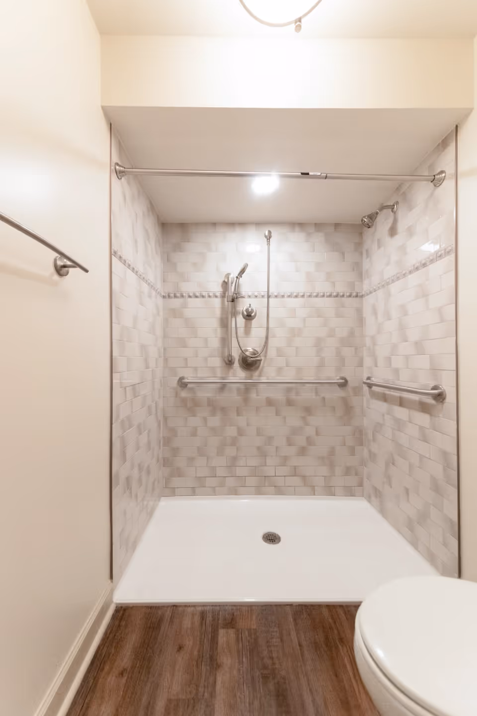 A clean, modern shower area with light gray subway tiles on the walls, a handheld showerhead, and two horizontal grab bars for support. The shower floor is white with a central drain, and the bathroom floor outside the shower is wood-patterned. A white toilet is partially visible on the right side.