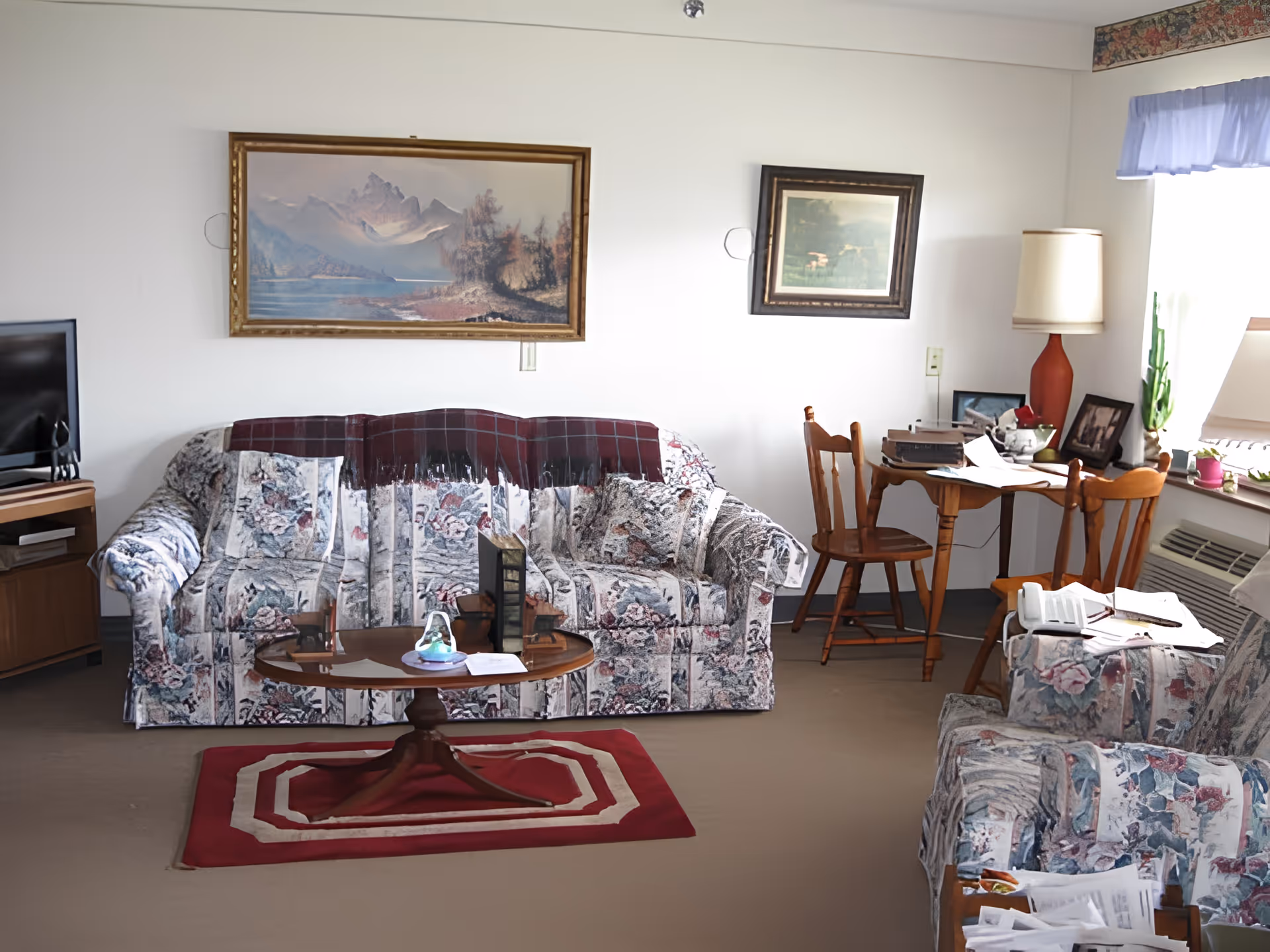 A cozy living room with floral patterned sofa and armchair, a wooden coffee table on a red rug, a small wooden desk with two chairs, a table lamp, and framed paintings on the white walls. A TV is placed on a wooden stand in the corner, and a window with a light blue valance lets in natural light.