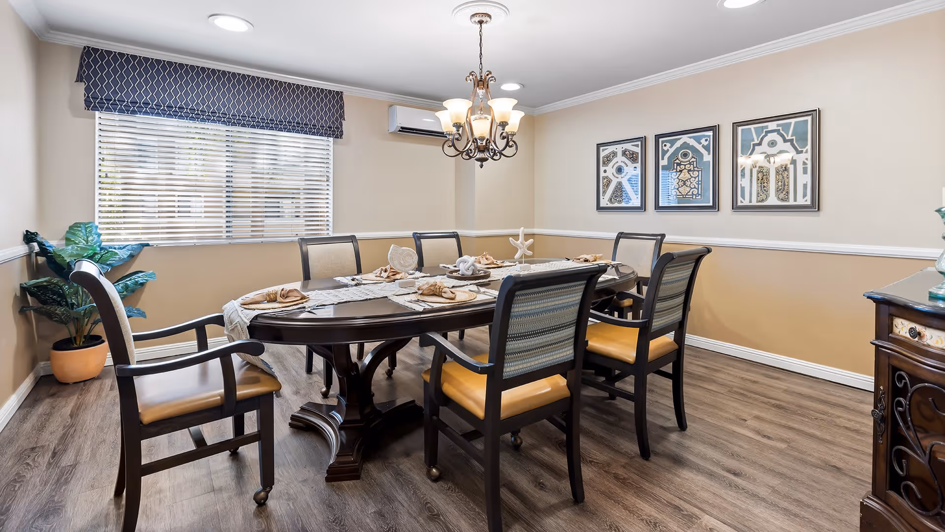 A dining room with a dark wooden oval table set for six with beige napkins and glassware. The room has light beige walls with white trim, a window with blinds and a dark valance, a chandelier hanging above the table, three framed artworks on the wall, a potted plant in the corner, and a wooden sideboard on the right.