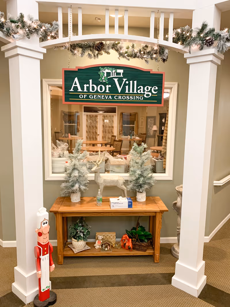 Interior entry display with a hanging "Arbor Village of Geneva Crossing" sign above a wooden table decorated with small frosted trees, a reindeer figure, and holiday accents.