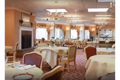A spacious dining room with round tables covered in white tablecloths, set with plates and glasses. The room features upholstered chairs, large windows with curtains, chandeliers, and a fireplace on the left side.