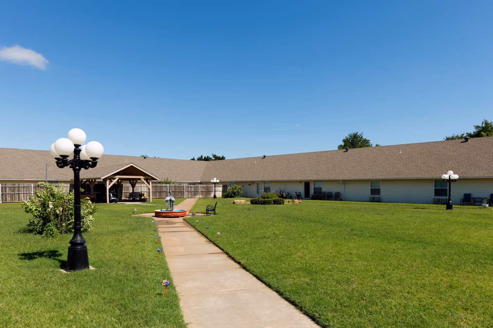 Outdoor courtyard area of Arbor House Of Mustang with a concrete walkway leading to a small fountain sculpture in the center. The courtyard is surrounded by a single-story building with a brown roof and white siding. There are black lamp posts with globe lights, benches, and a wooden gazebo in the background under a clear blue sky.
