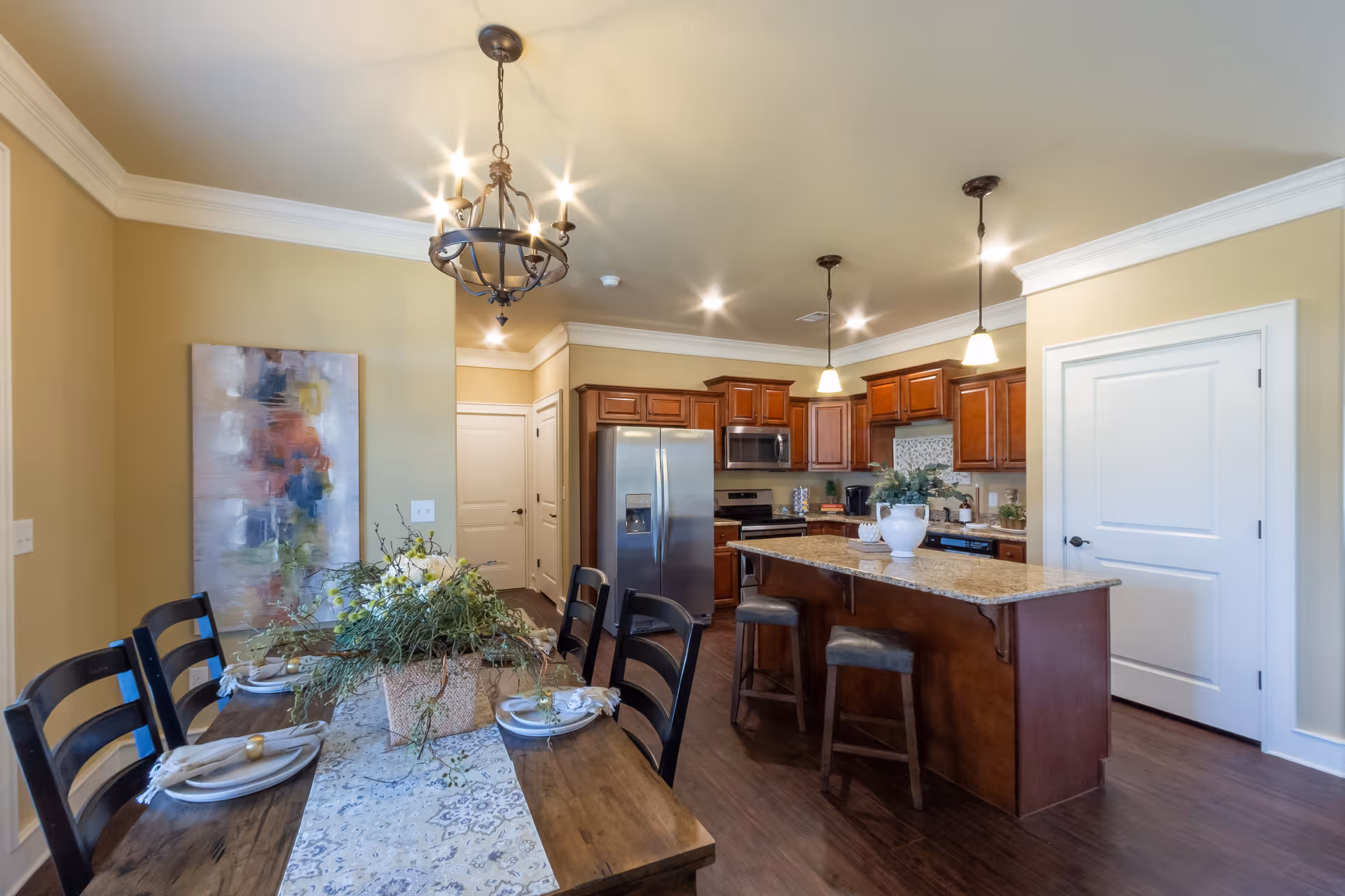 A bright and spacious kitchen and dining area featuring a wooden dining table set with plates and napkins, a floral centerpiece, a kitchen island with two bar stools, stainless steel refrigerator, microwave, and wooden cabinetry. The room is illuminated by ceiling lights and pendant lamps, with beige walls and dark hardwood flooring.