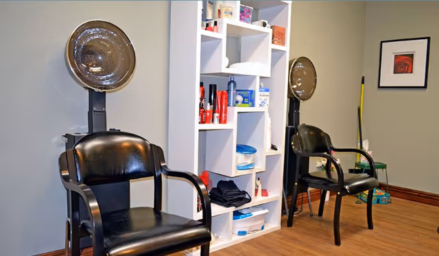 Interior view of a salon area with two black salon chairs positioned in front of hair dryers. Between the chairs is a white shelving unit stocked with various hair care products and supplies. The room has wooden flooring, gray walls, and a framed picture on the wall.