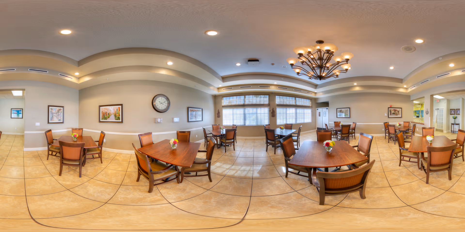 A spacious dining room with multiple wooden tables and chairs arranged neatly. Each table has a small floral centerpiece. The room features large windows with blinds, beige walls adorned with framed artwork, a large clock, and a decorative chandelier hanging from the ceiling. The floor is tiled with light-colored tiles.