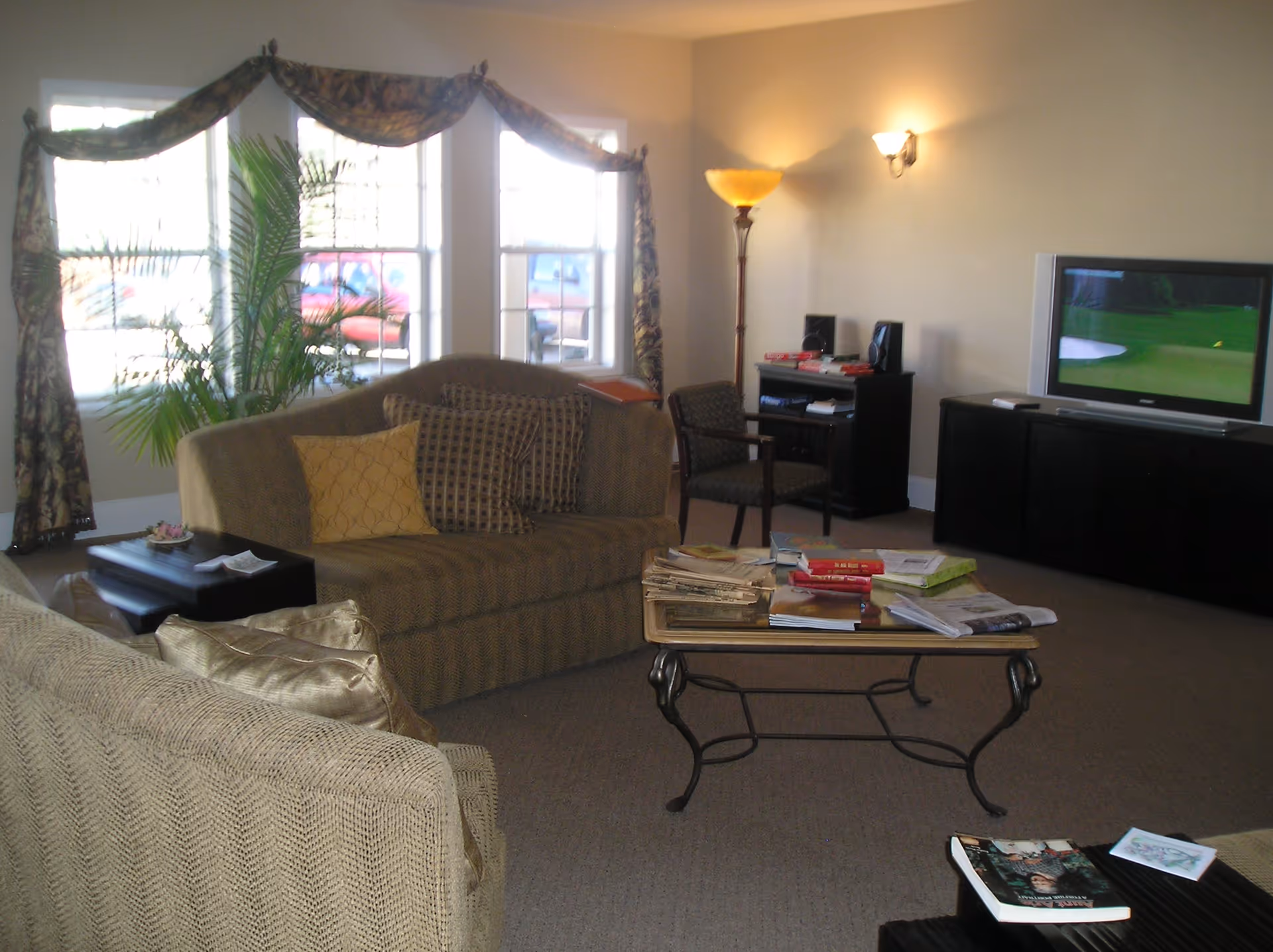 A cozy living room with two beige patterned sofas, a glass coffee table with magazines and books, a TV on a black stand showing a golf course, a floor lamp, a chair, and large windows with floral curtains letting in natural light.