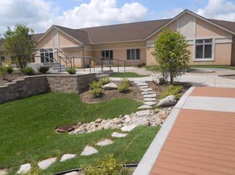 Front exterior of a single-story senior living building with landscaped lawn, stone stepping path, ramped entry, and blue sky.