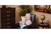 Cozy living room corner with a dark dresser, cushioned chair, pillows, plant, globe, and art.