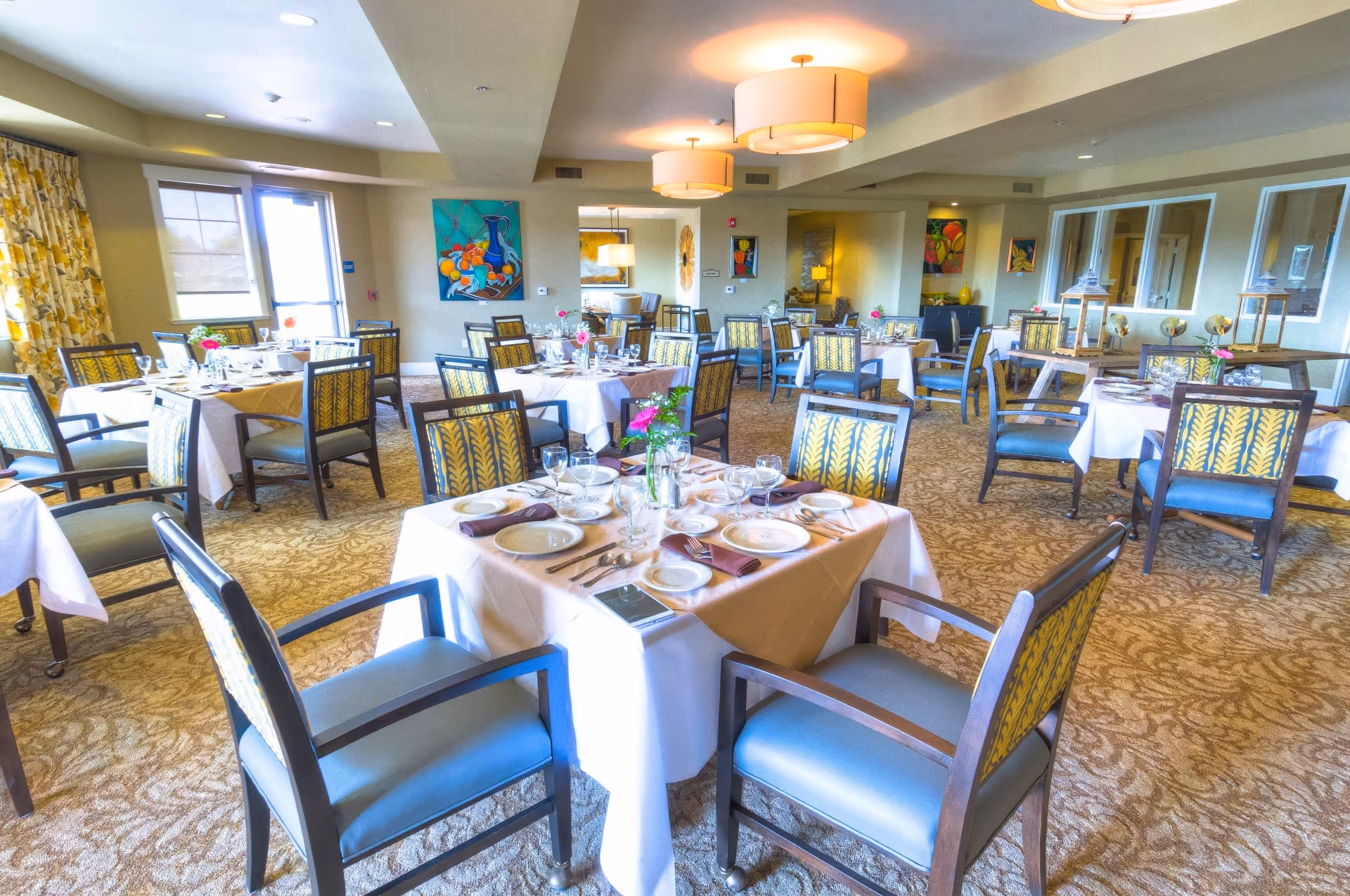 A bright and spacious dining room with multiple tables set with white tablecloths, plates, glasses, and cutlery. The chairs have patterned upholstery and the room is decorated with colorful paintings and floral arrangements. Large windows allow natural light to fill the space.
