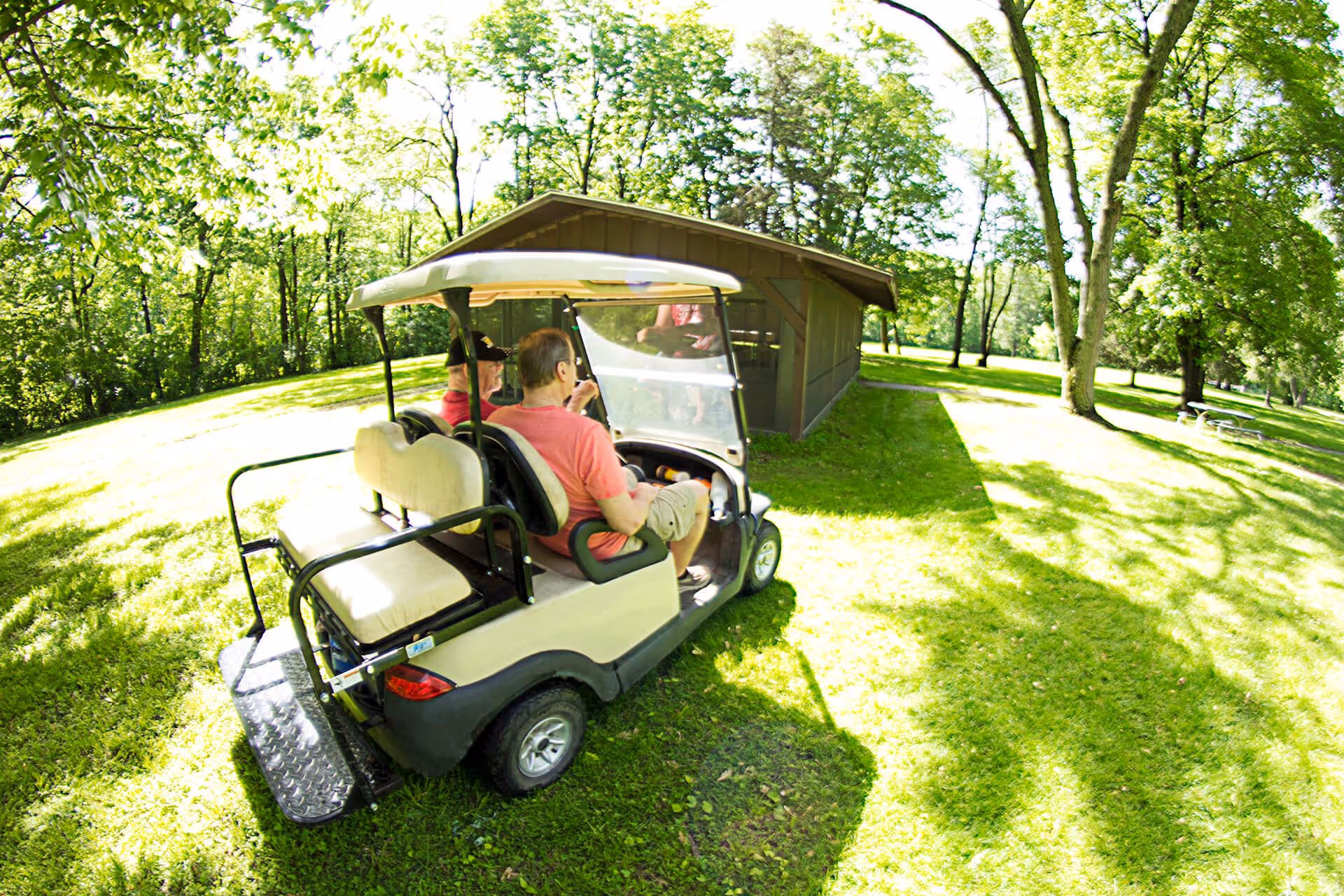Two elderly individuals riding in a golf cart on a grassy area surrounded by trees near a brown wooden building on a sunny day.