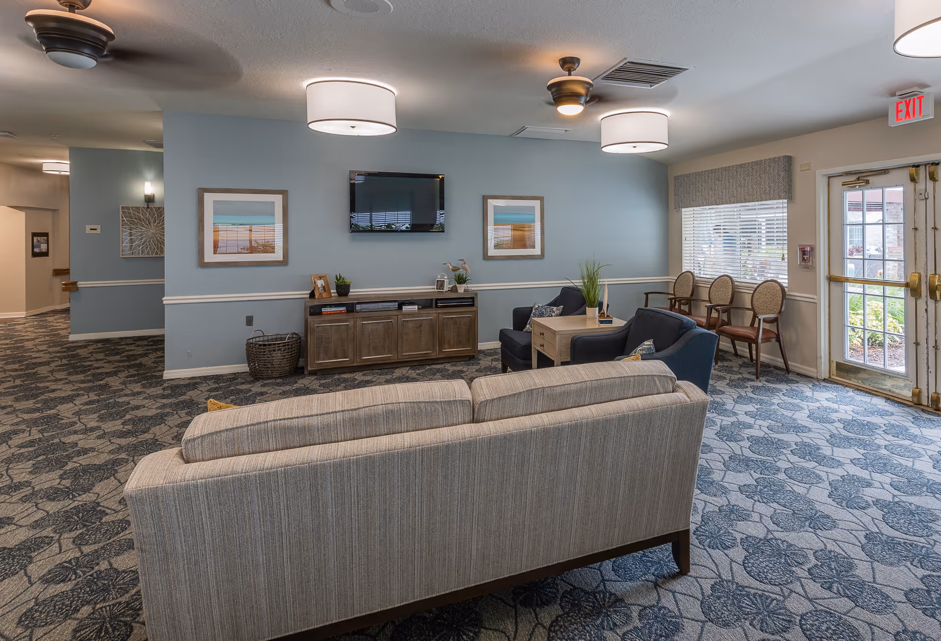 A cozy living room area with a striped beige sofa facing two dark blue armchairs and a wooden coffee table. The room has light blue walls adorned with two framed pictures and a flat-screen TV mounted between them. The floor is covered with a patterned carpet in shades of blue and gray. There are ceiling lights and fans, a window with blinds, and an exit door leading outside. Additional chairs are lined up against the wall near the window.