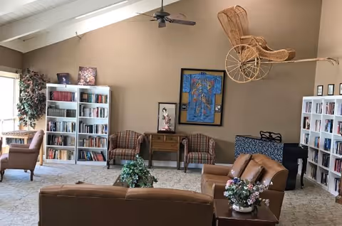 A cozy living room area in an assisted living community featuring brown leather sofas, two armchairs, bookshelves filled with books, a small wooden table with a plant, and a ceiling fan. The walls are decorated with framed artwork and a vintage wicker baby carriage mounted high on the wall.
