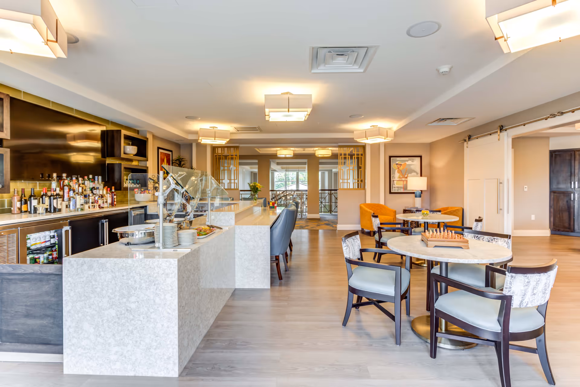 A modern dining and lounge area in Arbor Terrace Highland Park featuring a long marble countertop with a buffet setup, bottles of wine and liquor on shelves behind the counter, several round tables with chairs, a chessboard on one table, and comfortable seating with armchairs. The room is well-lit with ceiling lights and has a light wood floor.