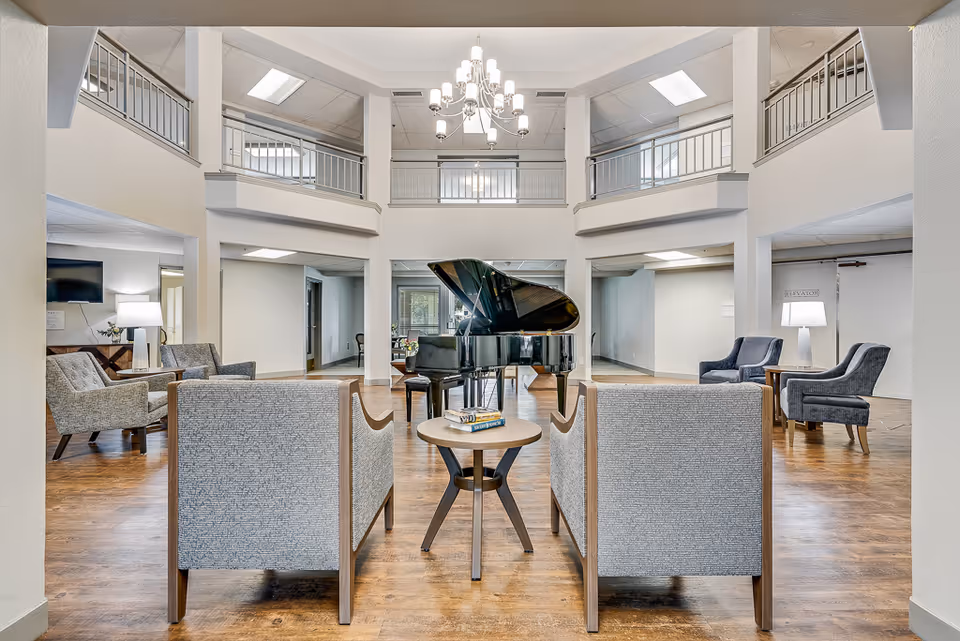 Bright two-story common lounge in a senior living facility featuring a grand piano, seating group, and chandelier.