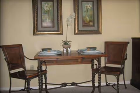 An indoor dining nook with a wooden table, two chairs, a plant centerpiece, and framed art.