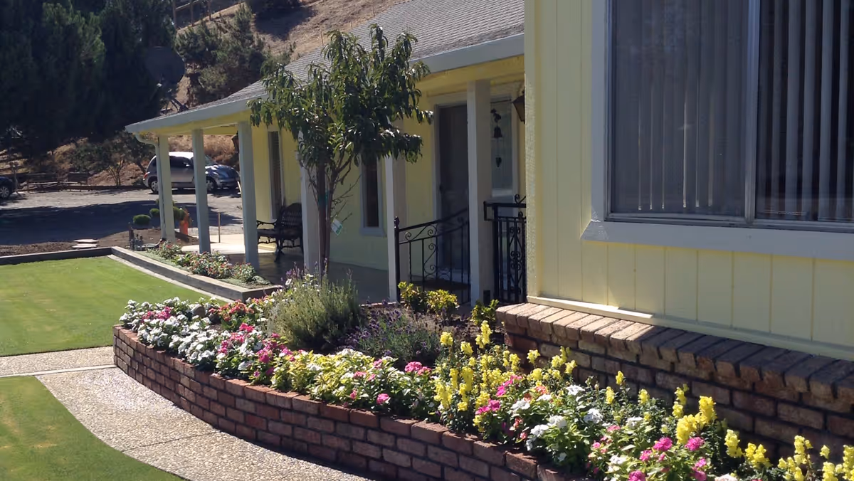 Front yard of a yellow house with a brick flowerbed, colorful flowers, green lawn, and a covered porch.