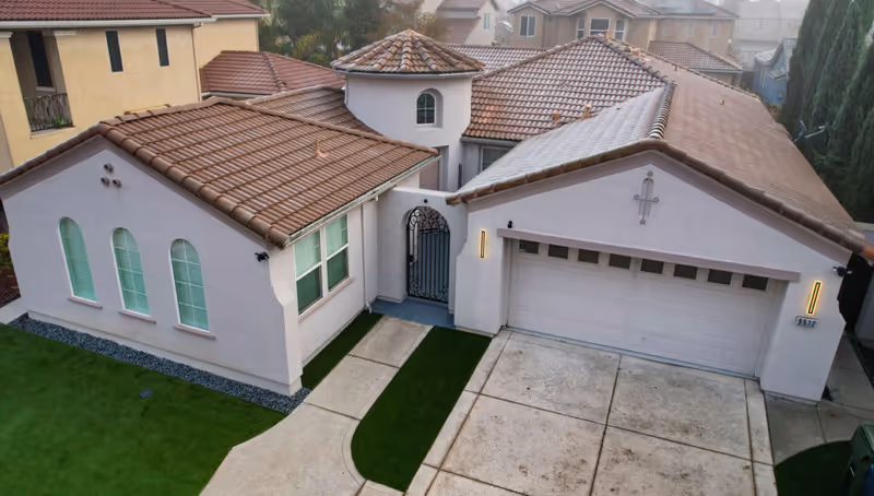A single-story residential building with a tiled roof and white exterior walls. The house features three arched windows on the left side, a gated entrance in the center, and a double garage door on the right. The driveway and walkway are concrete with a strip of artificial grass. The surrounding area includes neighboring houses and some tall trees on the right side.