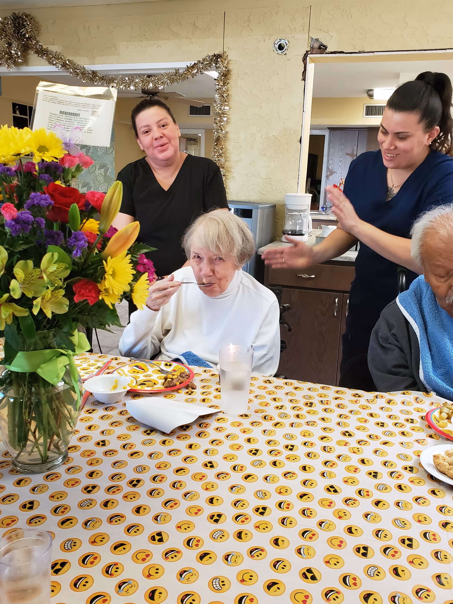 An elderly woman eats at a dining table with caregivers standing behind her, a vase of flowers on the table, and another resident seated nearby.