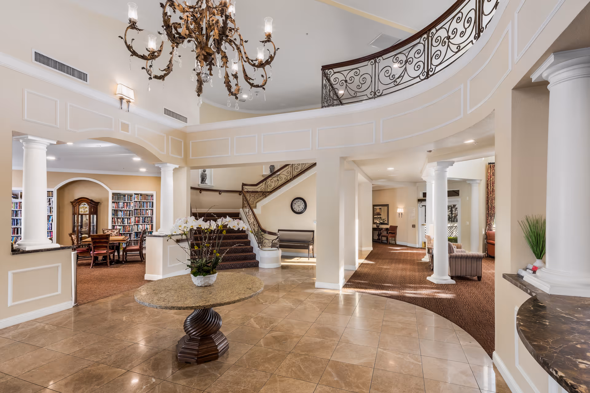 Spacious and elegant interior of a senior living facility featuring a large chandelier, a round table with a floral arrangement in the center, a staircase with ornate railing, and a cozy library area with bookshelves and a table with chairs.