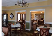Interior view of a dining room in a senior living facility with round tables covered with white tablecloths, wooden chairs, a chandelier hanging from the ceiling, and a china cabinet in the corner. The room has yellow walls with white trim and an adjoining room visible through two open doorways.