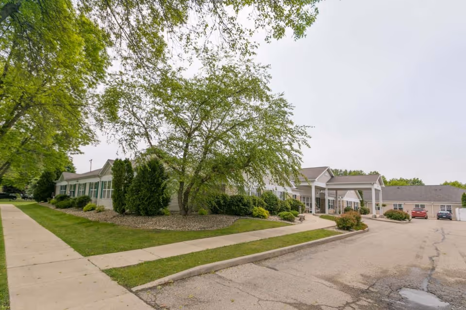 Exterior view of Madison Pointe Senior Living facility showing a single-story building with a covered entrance, surrounded by green trees, bushes, and a parking area with a few cars.