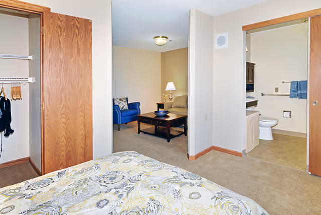 View from a bedroom showing part of a bed with patterned bedding, an open closet with hangers, a small living area with a blue armchair, beige sofa, wooden coffee table, and a lamp, and a bathroom with a toilet and towels visible.