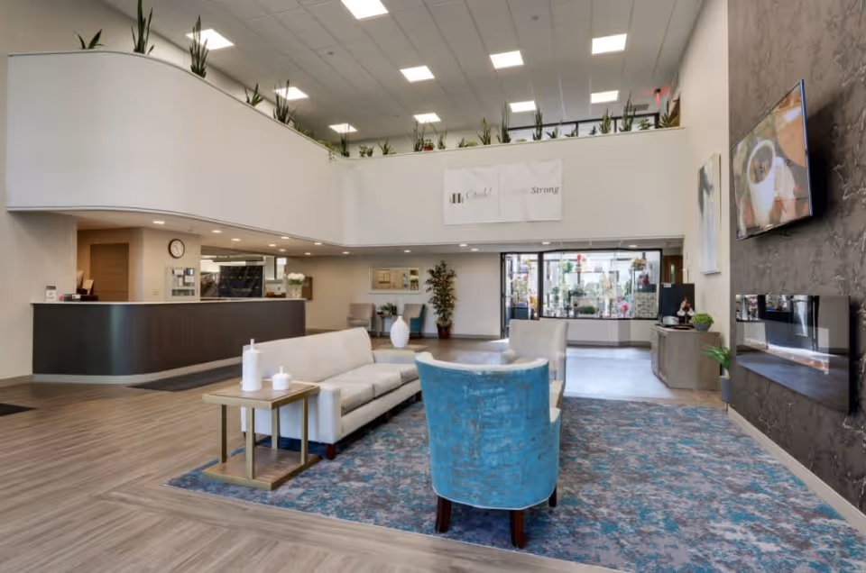 A spacious and modern senior living facility lobby with a reception desk on the left, a seating area with a white sofa and blue chairs on a patterned rug in the center, and a large flat-screen TV mounted on a textured wall to the right. The ceiling is high with recessed lighting, and there are plants along the upper balcony railing. A glass door entrance is visible in the background.