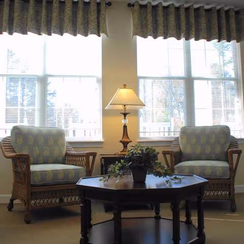 A cozy sitting area with two wicker armchairs featuring patterned cushions, a wooden coffee table with a potted plant, and a table lamp placed between the chairs in front of large windows with valance curtains.