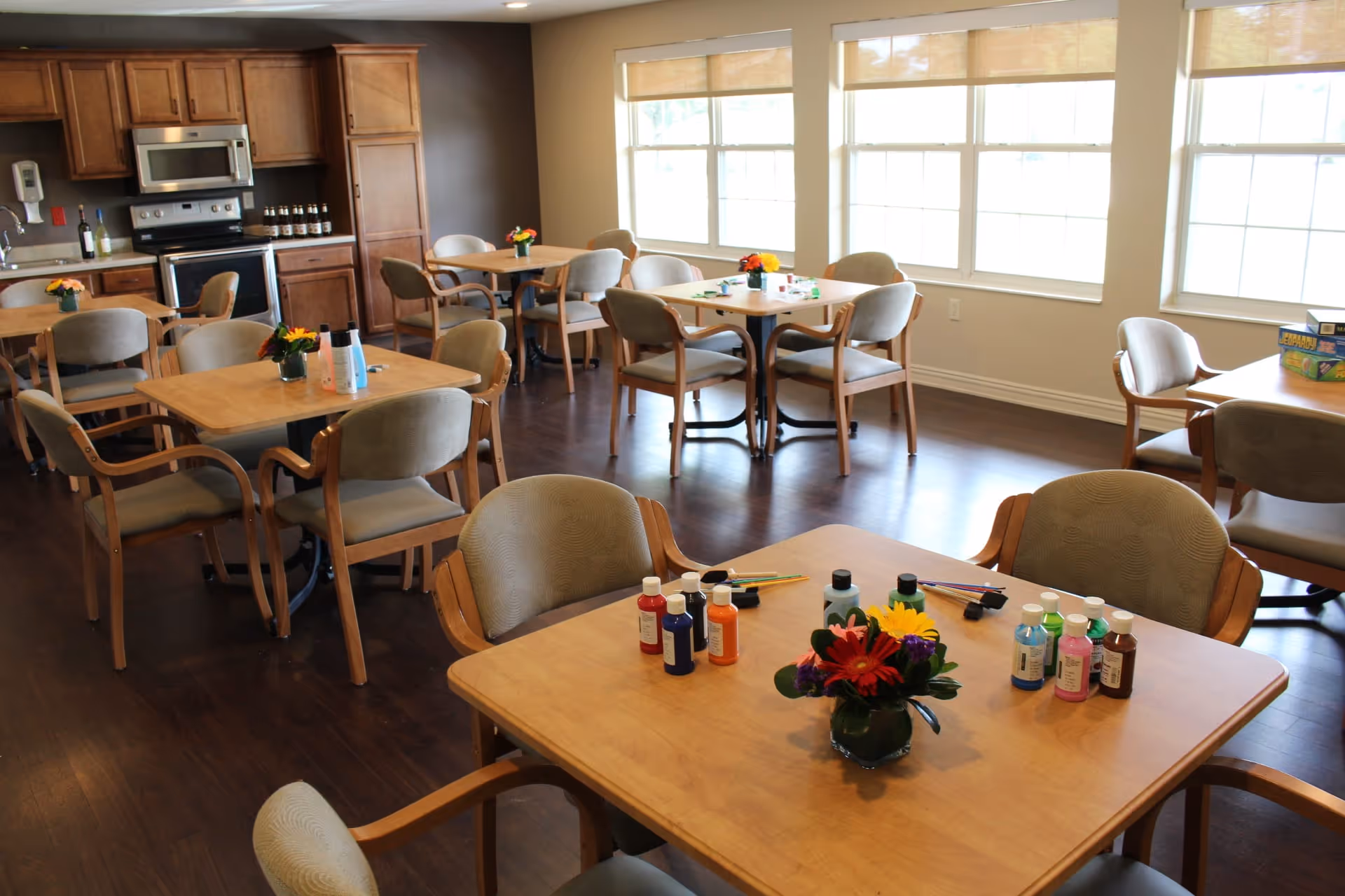 A bright room with several wooden tables and cushioned chairs arranged around them. Each table has a small flower arrangement and some have bottles of paint and paintbrushes, suggesting an arts and crafts activity area. The room has large windows letting in natural light and a kitchen area with wooden cabinets and stainless steel appliances in the background.