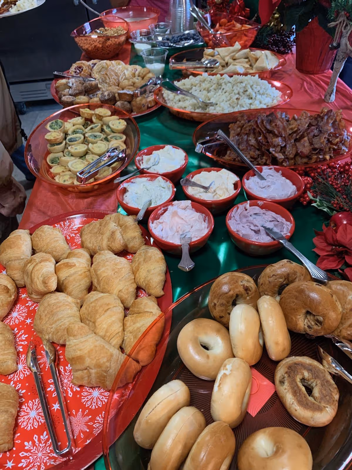 A buffet table with a variety of breakfast foods including croissants, bagels, assorted spreads in bowls, bacon, scrambled eggs, mini quiches, muffins, and fresh fruit, all arranged on red and green tablecloths with holiday decorations.
