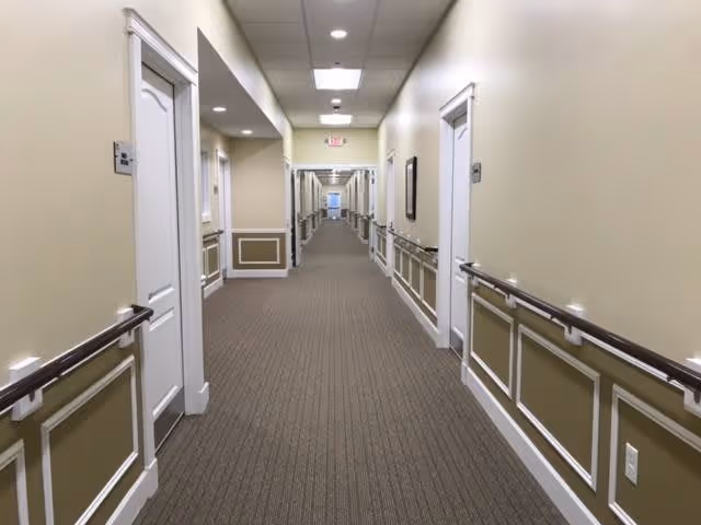 A long, well-lit hallway in a senior living facility with beige walls, white doors on both sides, handrails along the walls, and a carpeted floor. The ceiling has recessed lighting and an exit sign is visible at the far end.