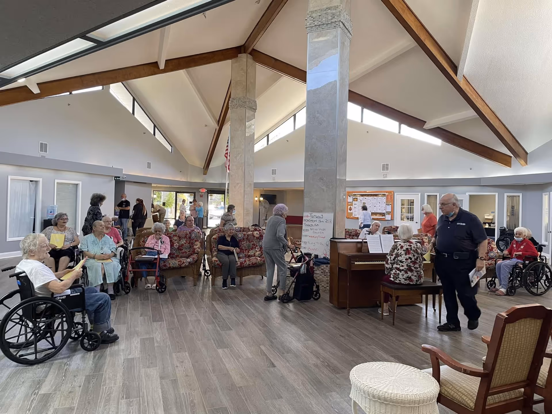 A spacious senior living facility common area with high vaulted ceilings and large windows. Several elderly residents, some in wheelchairs and others seated on floral-patterned sofas, are gathered around a piano where a woman is playing music. A man stands nearby holding a booklet. The room has wooden beams, light-colored walls, and wood flooring. A whiteboard with notes is visible near the piano.