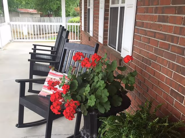 A porch area with black rocking chairs lined up against a brick wall. One chair has a striped cushion. There is a black planter with red flowers and green leaves in the foreground, and a white railing along the edge of the porch.