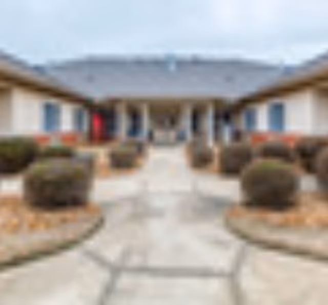 Outdoor courtyard area of a senior living facility with a paved walkway, trimmed bushes, and single-story buildings on either side under a cloudy sky.