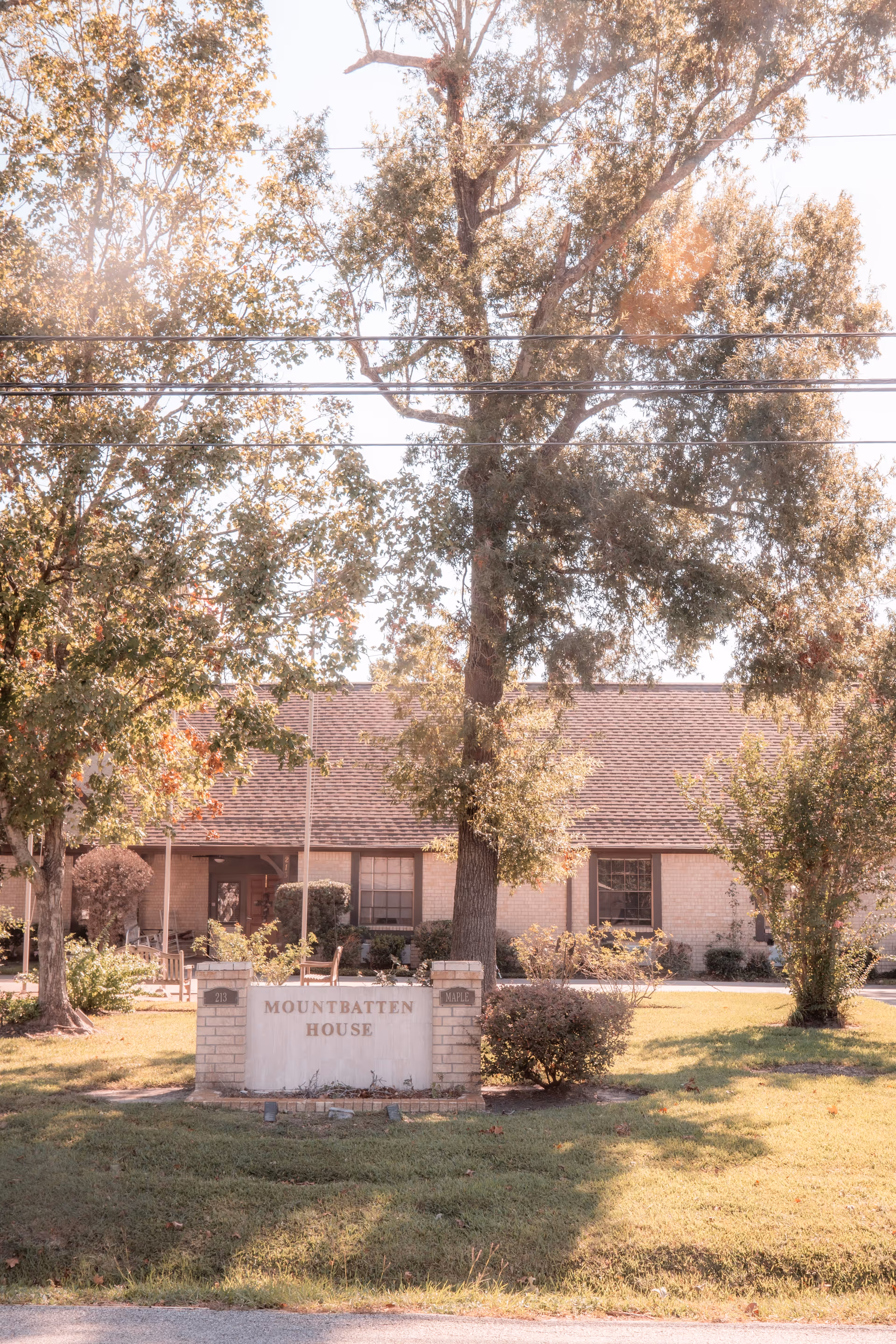 Exterior view of Mountbatten House, a single-story brick building with a shingled roof, surrounded by trees and greenery. A sign in front reads 'Mountbatten House' with the numbers 213 and the word 'Maple' on either side. The scene is lit by soft sunlight.