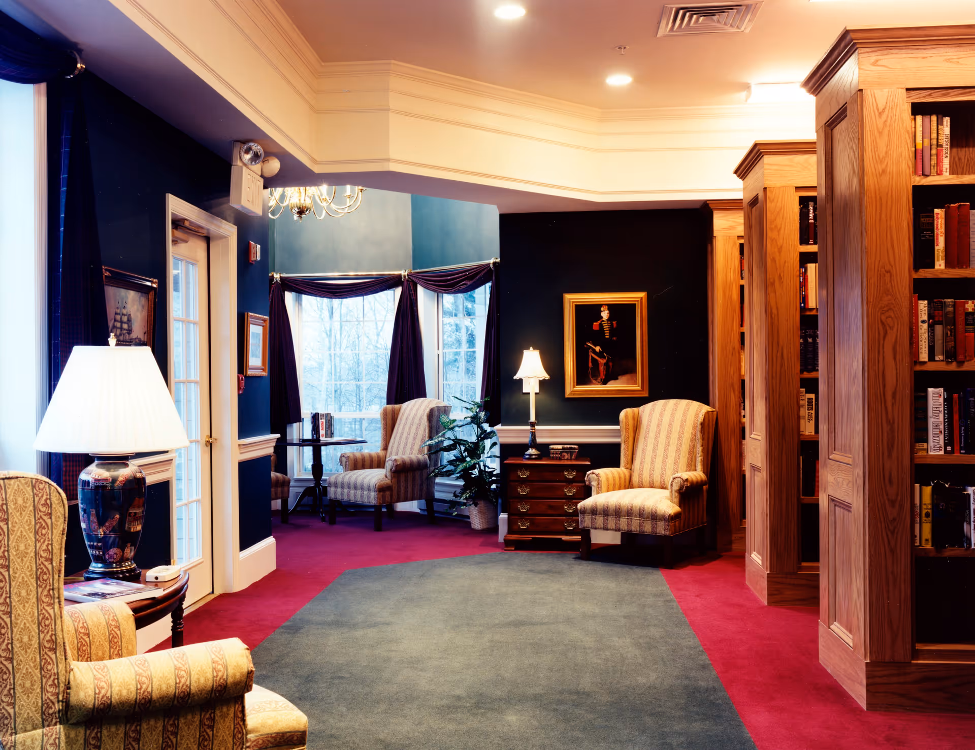 A cozy living room area in an assisted living facility featuring upholstered armchairs, wooden bookshelves filled with books, a side table with a lamp, framed artwork on the walls, large windows with dark curtains, and a carpeted floor with a red border and blue center.