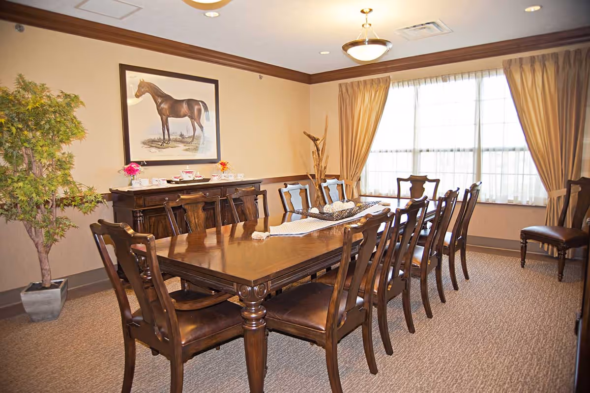 A well-lit dining room with a long wooden table surrounded by ten matching wooden chairs. The room features beige walls with a large framed picture of a horse on one wall, a sideboard with tea sets and flowers, a large window with beige curtains, and a potted plant in the corner.
