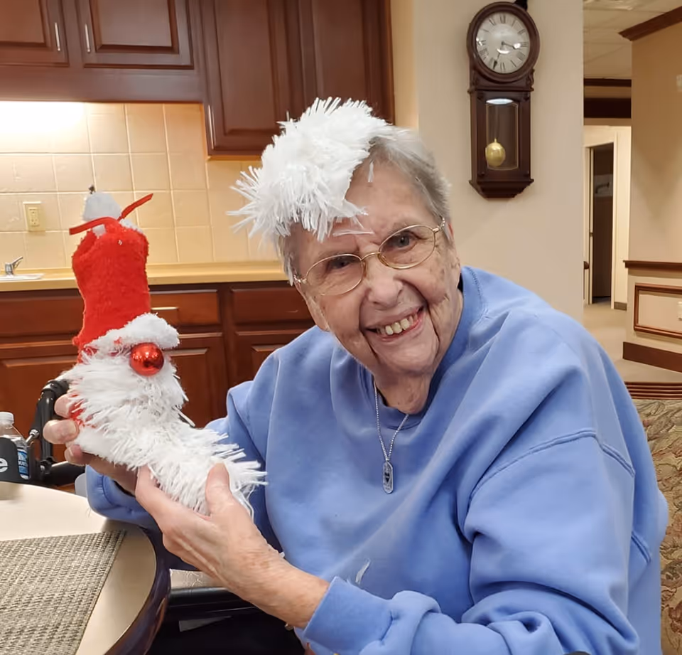 An elderly woman wearing glasses and a light blue sweatshirt is smiling and holding a festive Christmas decoration shaped like a white boot with a red Santa hat. She has a white fluffy decoration on her head. The background shows a kitchen area with wooden cabinets and a wall clock.